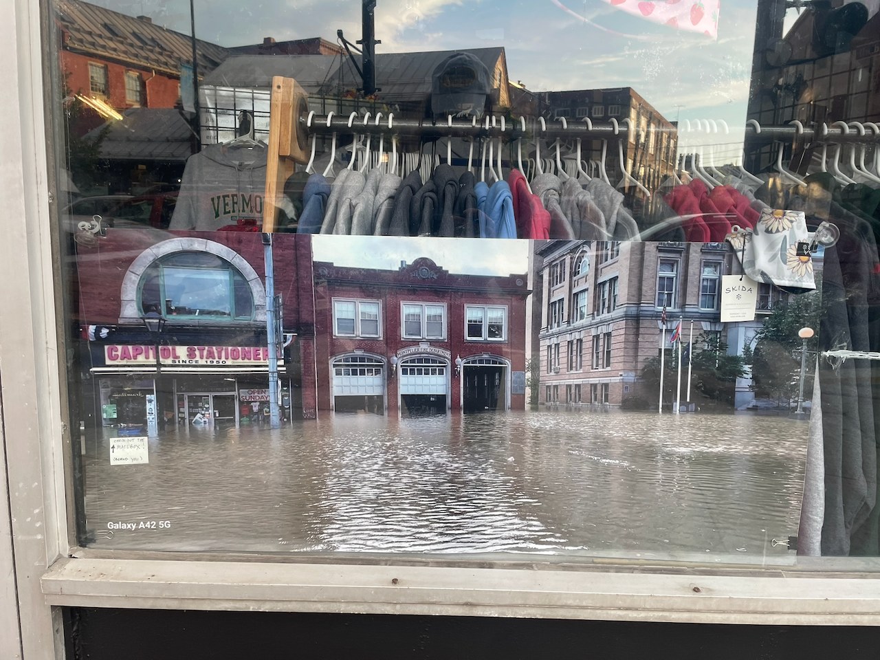 A print of downtown Montpelier under brown floodwater, displayed in a shop window with the present-day street reflected in the glass.
