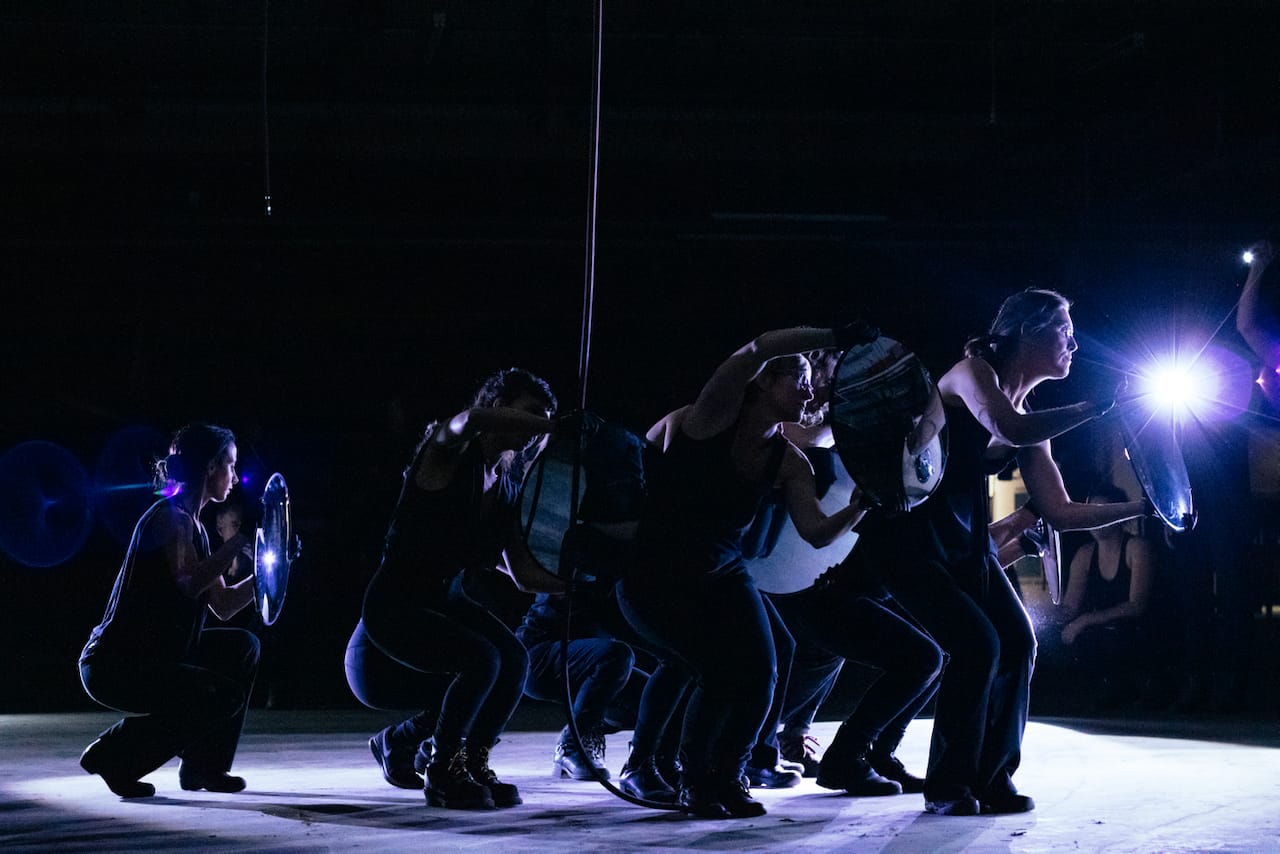 A performance ensemble caught in a shaft of light on a dark floor.